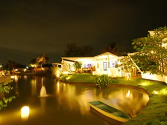 สวนริมน้ำยามค่ำ มีเรือไม้จอดริมตลิ่งและน้ำพุกลางสระ